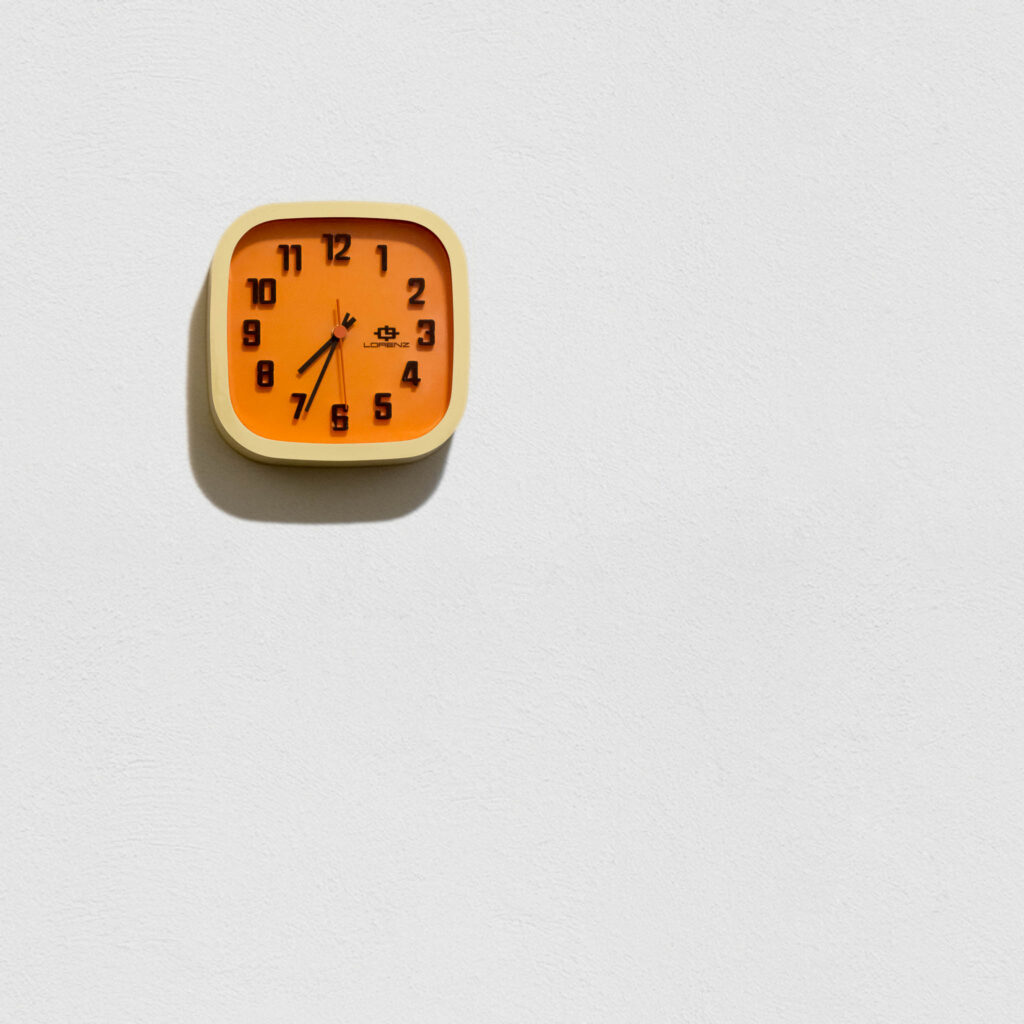 Un cadran d'horloge analogique orange, avec des aiguilles noires, sur un mur entièrement blanc — mais texturé, quand même. Il indique une heure bien précise: 7h34:29. Impossible de savoir s'il est matin ou soir, puisque la source de lumière est manifestement artificielle.  
